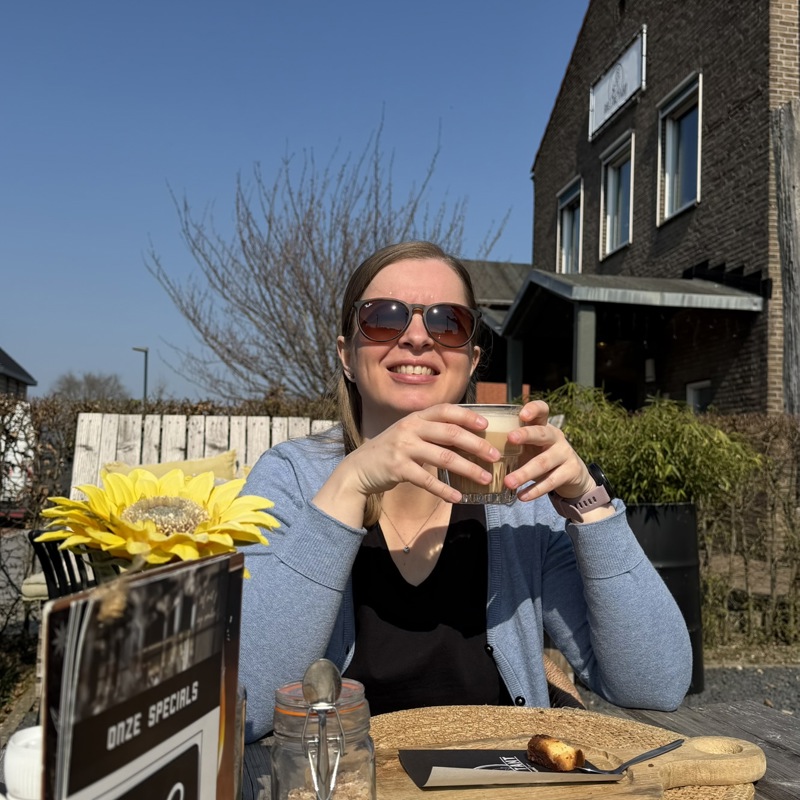 Vrouw drinkt een kopje koffie in de zon op een terras, met een strak blauwe lucht. 