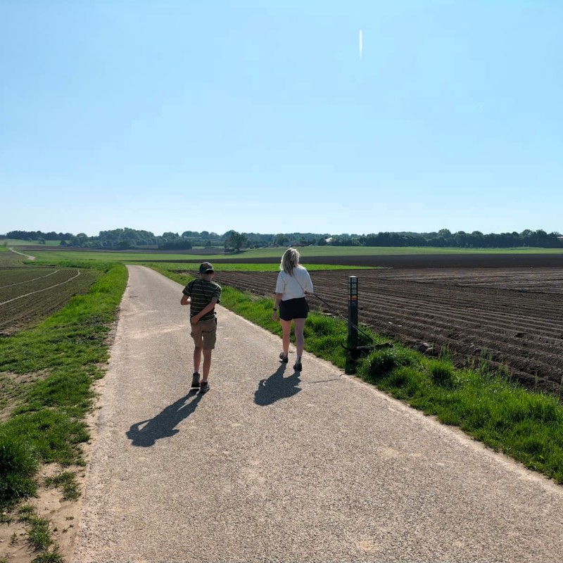 Moeder en zoon wandelen op een geasfalteerd pad met links en rechts landbouw. 