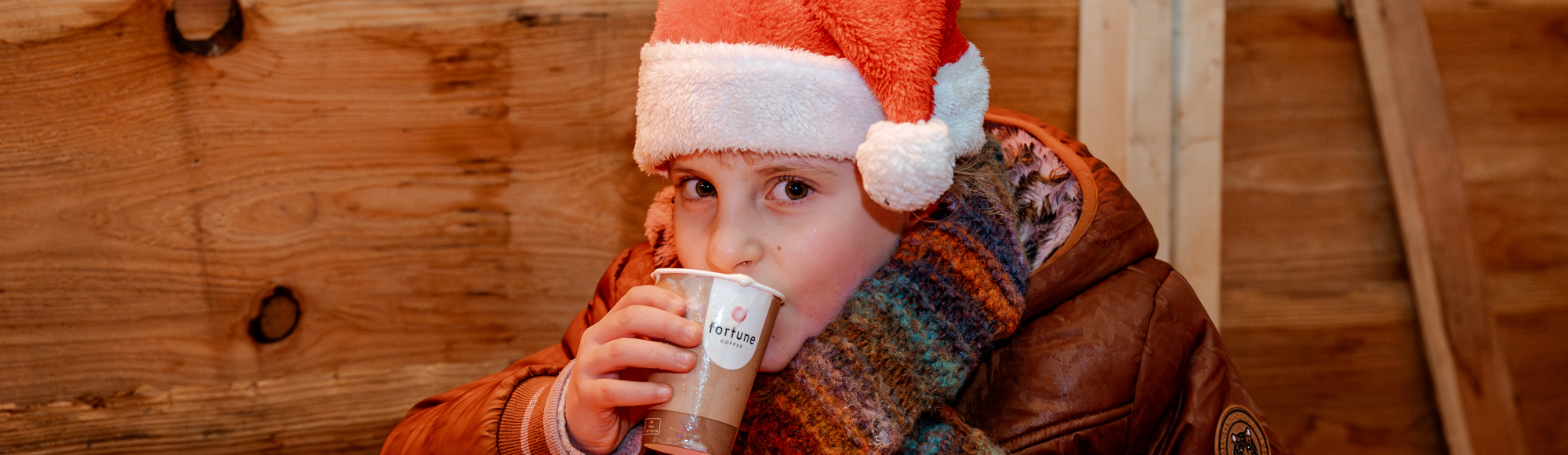 Kind met kerstmuts en winterjas drinkt iets in houten hut