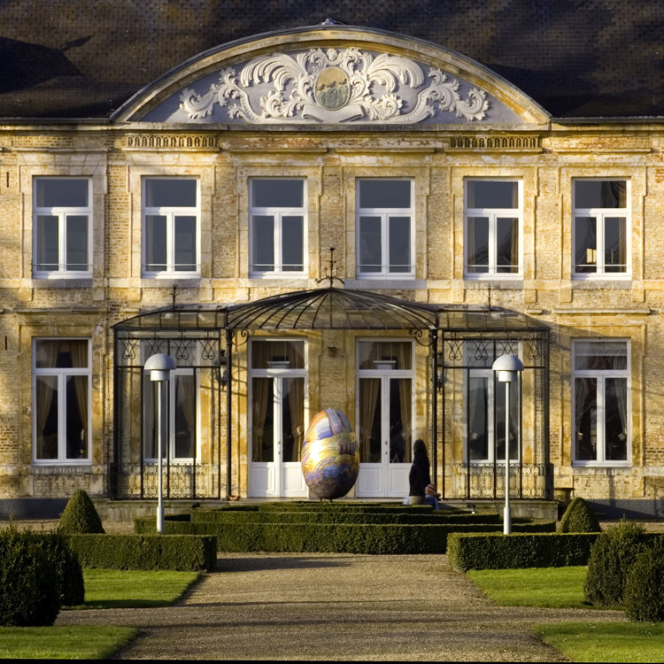 Vooraanzicht van een chateau met tuin en kunstwerk ervoor.