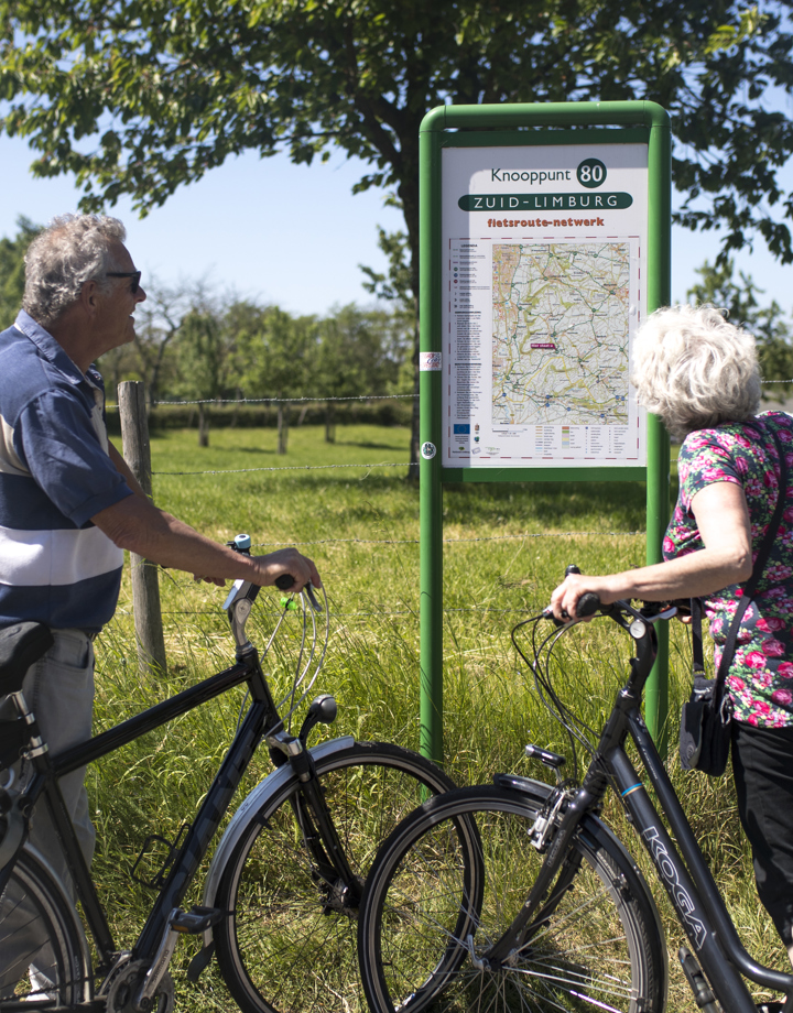Twee ouderen mensen met fiets in hun hand kijken naar een infopaneel met fietsknooppunten