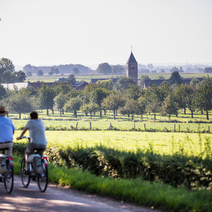 Koppel fietst langs weiland met uitzicht op de kerk en het dorpje Mesch