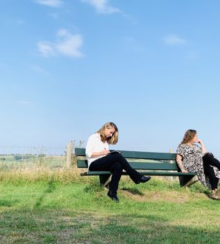 Jenneke en Celine zitten op een bankje met 1,5 meter afstand met op de achtergrond de zuid-limburgse heuvels