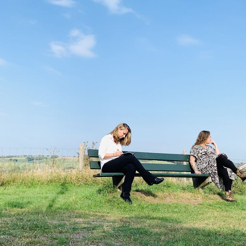 Jenneke en Celine zitten op een bankje met 1,5 meter afstand met op de achtergrond de zuid-limburgse heuvels