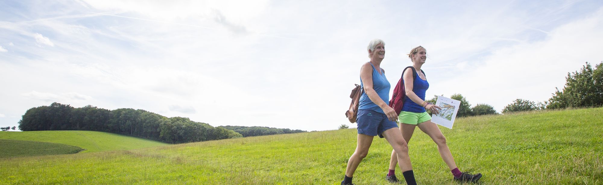 Twee vrouwen wandelen lachend door het landschap van Zuid-Limburg op een lichtbewolkte dag