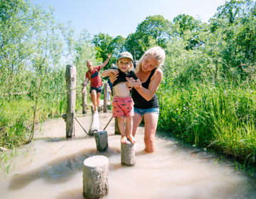 Moeder helpt haar kind met het lopen over de boomstronken in het water bij het blotevoetenpad