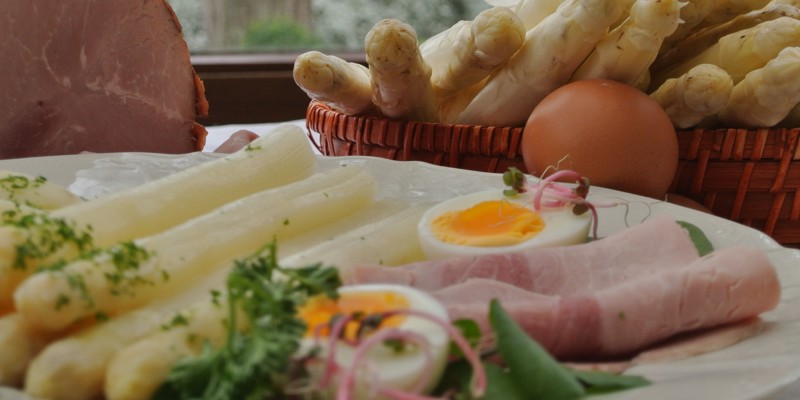Een bord met gekookte asperges, ham en ei, met op de achtergrond een mandje vol rauwe asperges