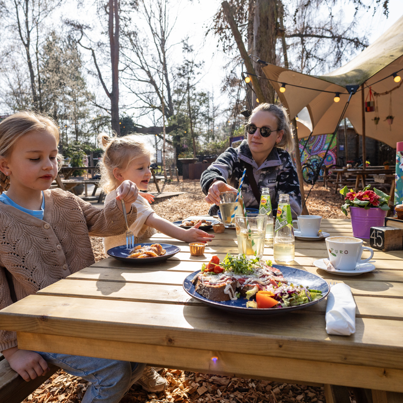 Moeder met twee kinderen zit aan houten picknickbank in horecagelegenheid met lunch op tafel.
