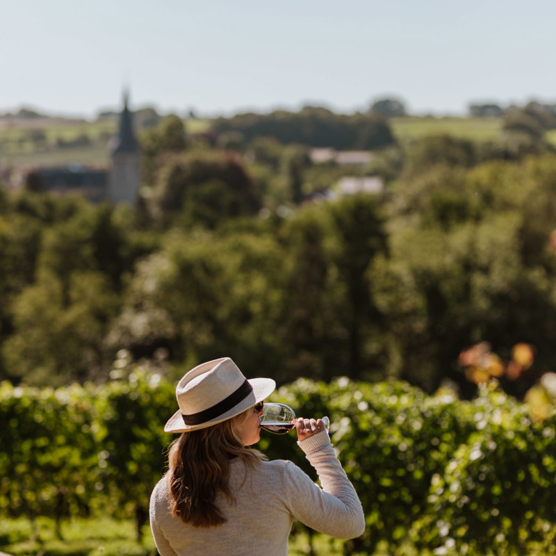 Een meisje zit op een muurtje en drinkt een rood glas wijn terwijl ze uitkijkt over de wijngaarden en de kerk van Noorbeek