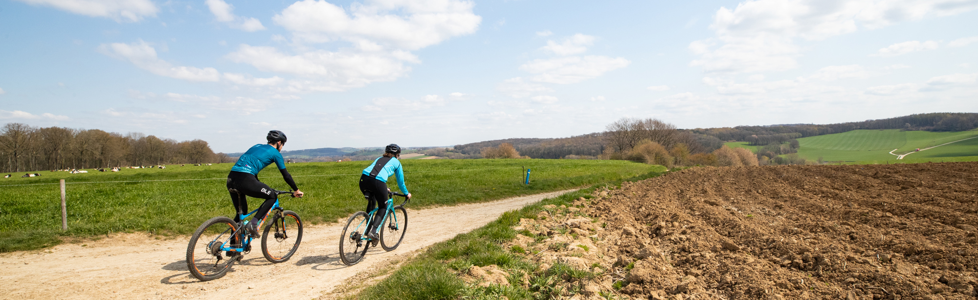 Man en een vrouw in blauw-zwarte fietstenues op gravelbikes op onverharde weg tussen de velden in het Heuvelland op een zonnige dag