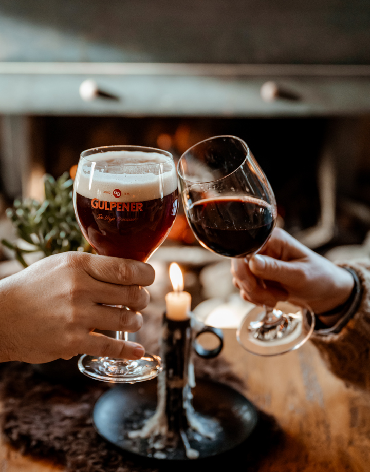 Bierglas en wijnglas proosten met aangestoken kaars in de achtergrond