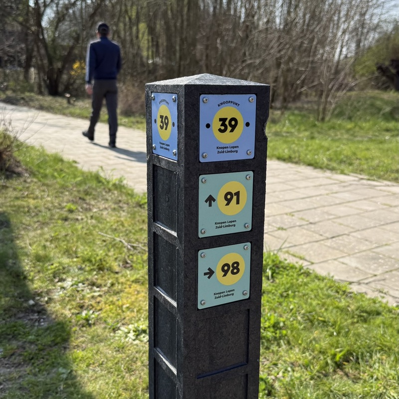 Close-up van een wandelknooppuntenpaal in de natuur, met een wandelaar op de achtergrond.