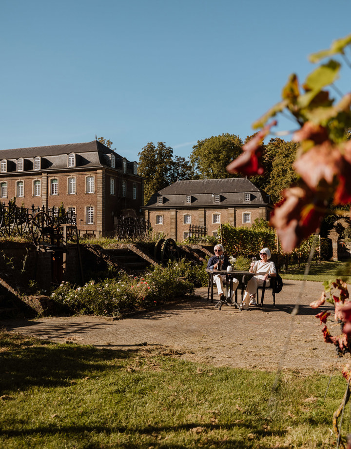 Een ouder koppel geniet aan een tafeltje van een glaasje witte wijn voor de wijngaard van Abdij Rolduc
