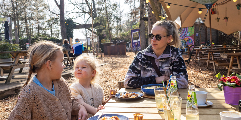 Moeder met twee kinderen zit aan houten picknickbank in horecagelegenheid met lunch op tafel.