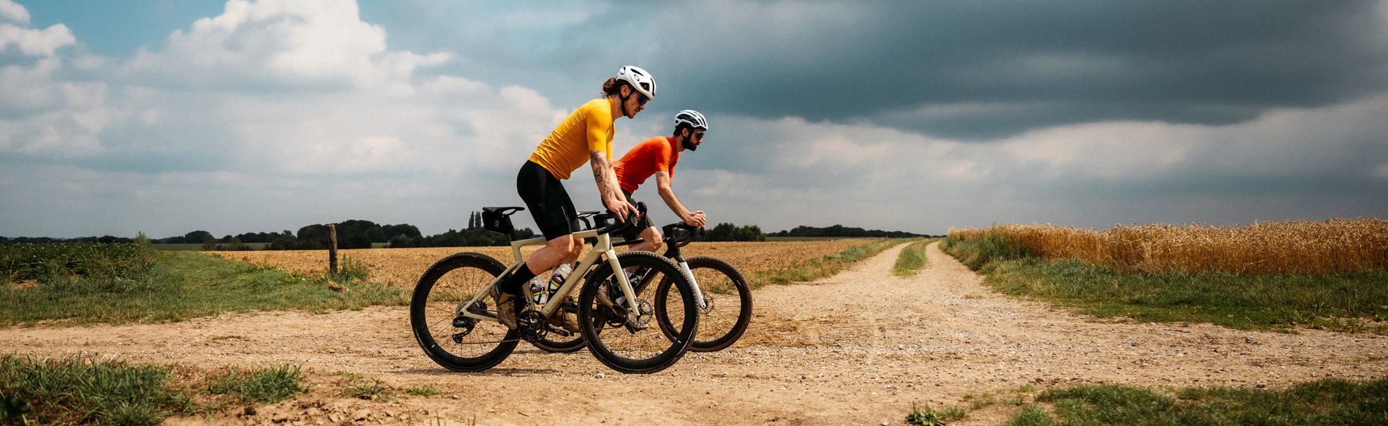 2 mannen met oranje fietspakjes op gravelbikes op onverharde weg tussen de velden