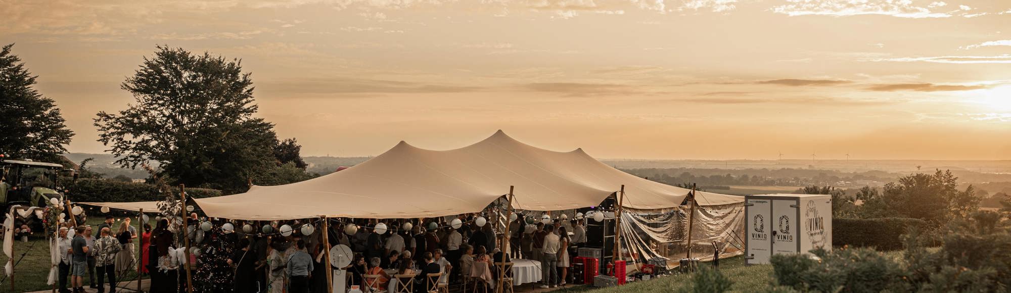 Mensen onder een feesttent met panoramauitzicht over het heuvelland bij zonsondergang