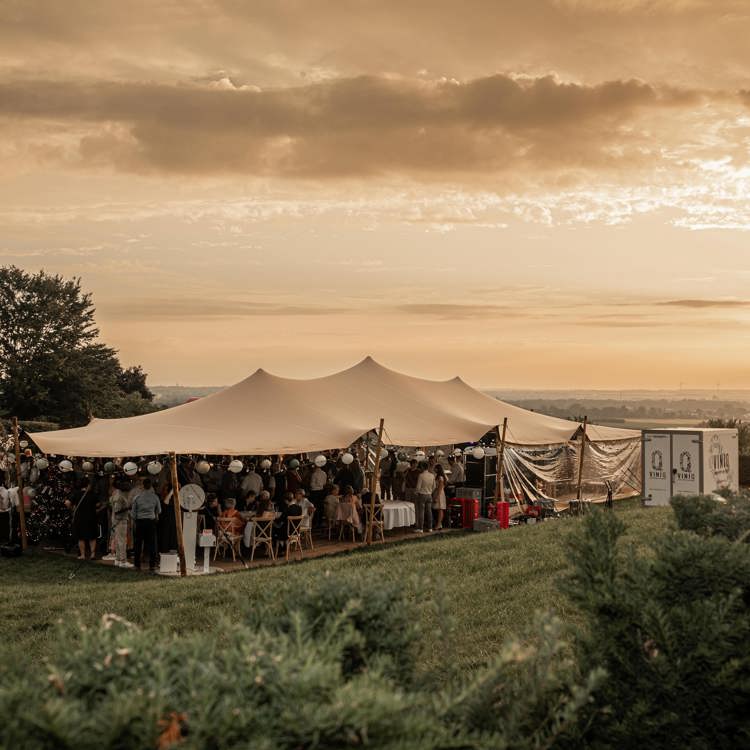 Mensen onder een feesttent met panoramauitzicht over het heuvelland bij zonsondergang