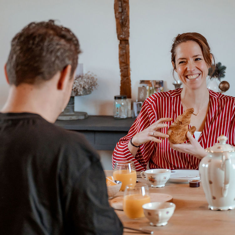 Koppel zit aan ontbijttafel en vrouw in badjas houdt lachend croissant vast.