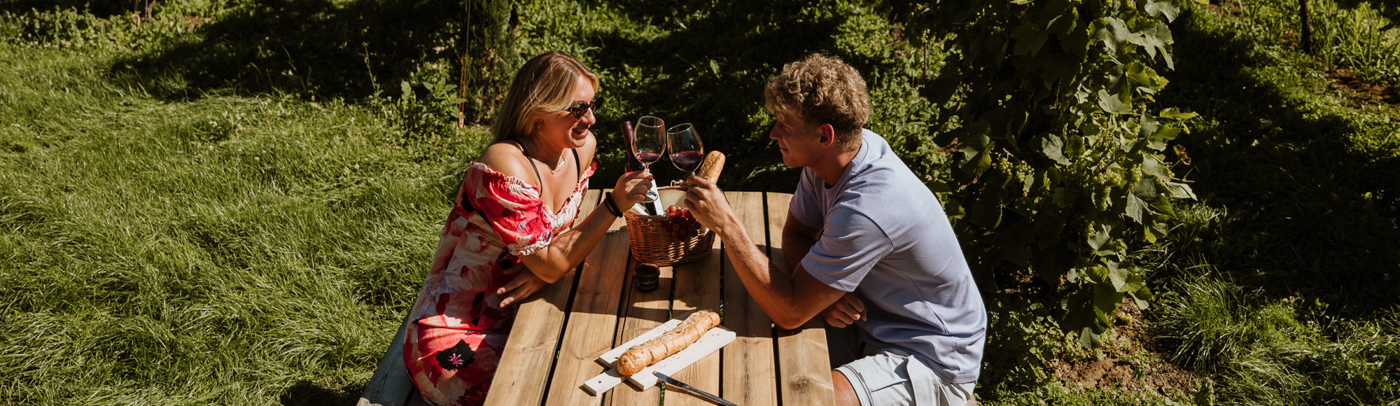 Een koppel proost met rode wijntjes terwijl ze zitten aan een picknickbank voor een wijngaard in Cadier en Keer
