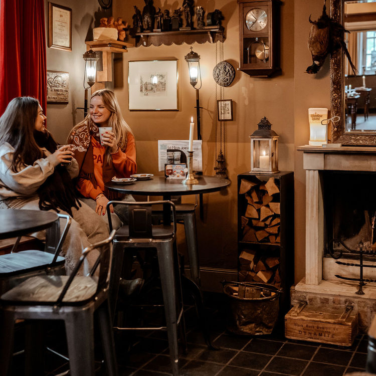 Twee vrouwen zitten aan een tafel met een warme drank bij een open haard en dragen winterkleding met sjaal en trui