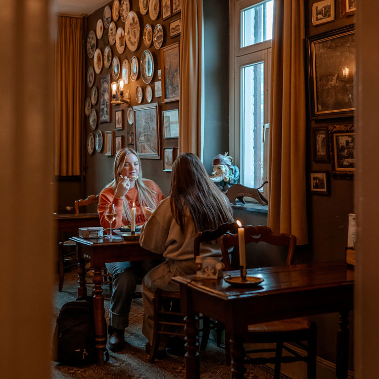 Twee vriendinnen zitten aan tafel in gezellig restaurant met oude schilderijtjes aan de muur. 