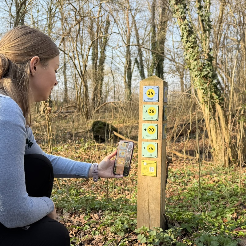 Wandelaar scant met haar telefoon de QR-code op een knopen lopen paaltje in de natuur.