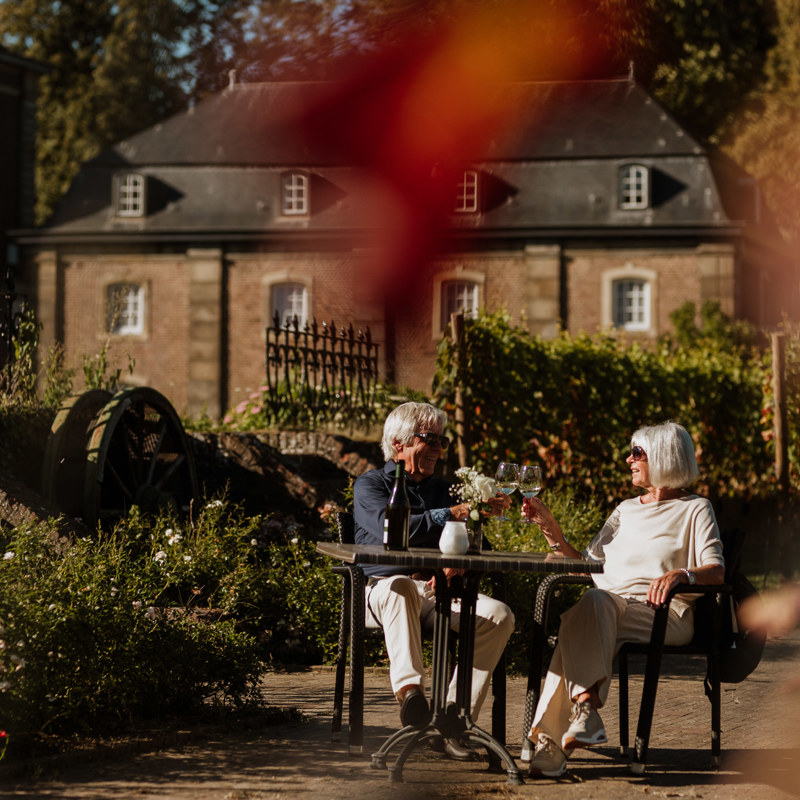 Een man en vrouw proosten aan een tafeltje voor de wijngaard van Abdij Rolduc